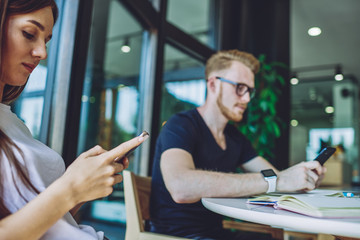 Millennial male and female friend typing text messages via mobile application using free wireless connection on cellular phones, caucasian friends searching information about language courses online