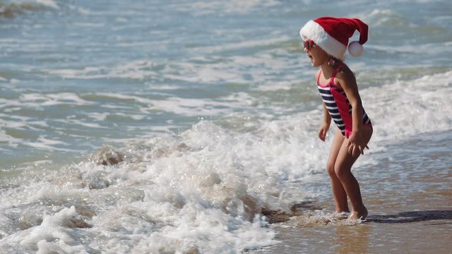 funny girl wearing swimsuit and red santa hat with sunglasses walks jumps on sand beach at rolling ocean waves slow motion