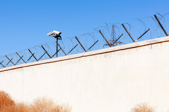 Security camera behind barbed wire fence stretched around prison walls. Security, surveillance camera watching area