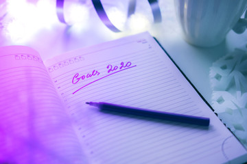 2020 goals written on a diary with light, hot coffe or tea and decoration. Happy New Year and motivation consept. Neon background