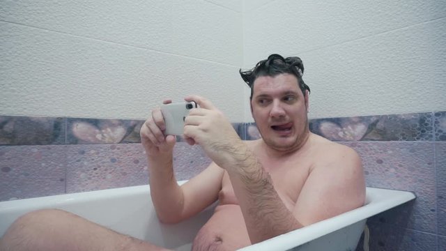 having fun at home.modern weird emotional guy lying in bath uses smartphone