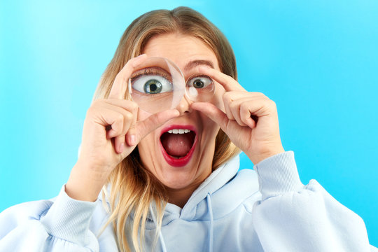 Fototapeta Young beautiful woman looking at the camera through a magnifier with a funny surprised expression, closeup isolated on pink. Enlarged eye, optical illusion