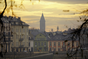 Obraz premium Fantastic Dramatic Bright sunset over old town through branches of trees, foggy weather. Selective Focus. Red, pink and blue bright sunset over city. Peaks of houses, antennas and chimneys on roofs.