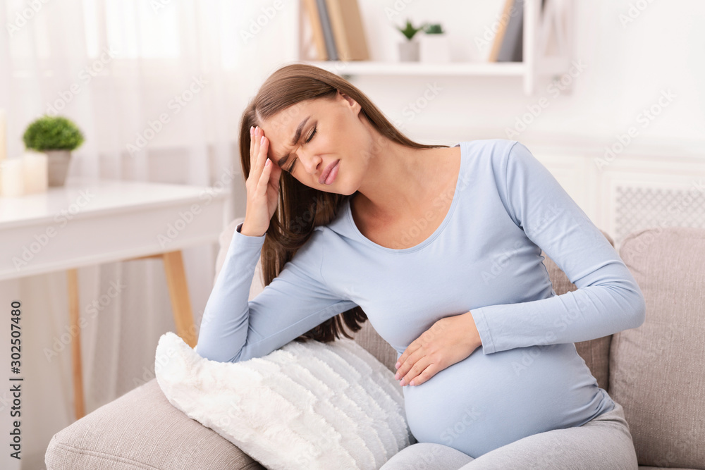 Wall mural pregnant lady having headache sitting on couch indoor