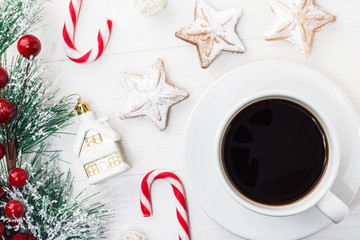 A white mug of black Americano coffee with Spruce branch tree on a white wooden background next to Christmas cookies, a candy cane and New Year's toys. Copy space. Top view.