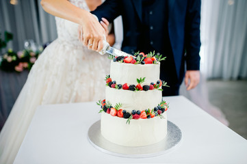 wedding ceremony. Hands of newlyweds cut a white three-tiered cake with strawberries and...