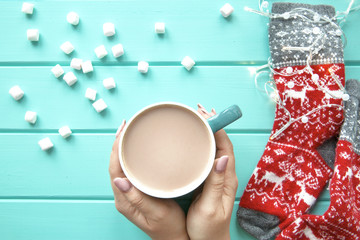 Hands holding a cup of tasty cocoa with marshmallow, festive lights and red woolen socks on wooden background, top view. 