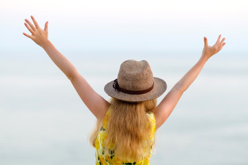 Beautiful little blonde girl with long hair in a wicker hat stands with her back, arms raised up, looks at the sea, summer, joy, delight, emotion