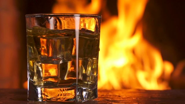 Ice cubes fall into a glass with whiskey, scotch or bourbon standing on a table the background of a fireplace with a flame. Alcoholic drink concept. Fire background. Drink alone. Close up Slow motion.