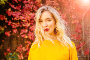 Lifestyle portrait of happy blond woman in yellow sweater with red lips flying hair, windy autumn sunny day on red brick wall with climbing grapes on background. Close up portrait.