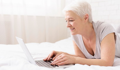 Senior lady browsing on laptop, resting on bed