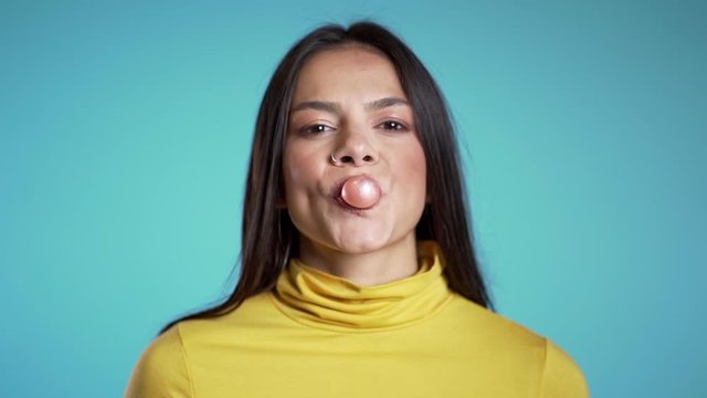 Playful girl in yellow wear blows bubblegum, chewing gum in slow motion. Pretty hispanic woman, piercing stands on blue background.