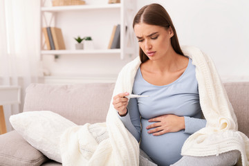 Obraz na płótnie Canvas Pregnant Girl Holding Thermometer Sitting On Sofa At Home