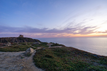 Wide-angle panorama of a beautiful lilac sunset overlooking the ocean with rocks in the foreground and a path leading up to a bunker.