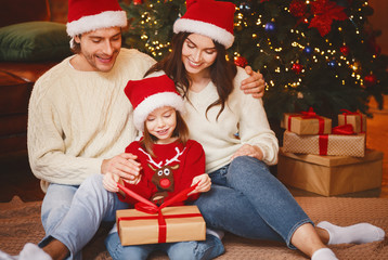Obraz na płótnie Canvas Cute Christmas family sitting next to festive tree, opening gifts