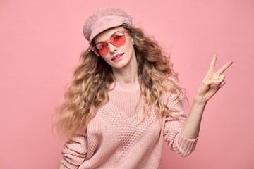 Easy-going Woman happy smiling in Stylish fashion pink colored outfit. Beautiful young Girl in Trendy jumper, make up. Joyful blonde lady shows peace sign in pink cap, fashionable happiness concept