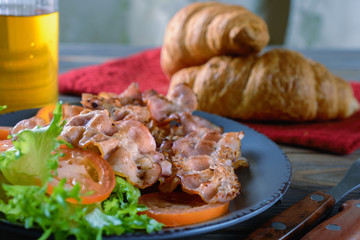 Hot fried bacon on green lettuce leaves and sliced ​​tomato on a plate next to croissants and a glass of juice are ready for breakfast or lunch