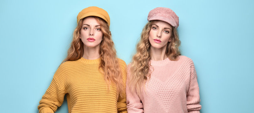 Fashionable woman in stylish outfit, makeup posing on blue. Two Beautiful blonde redhead girl, trendy pink yellow jumper, cap, fashion hair. Adorable sister friend, colorful pastel concept
