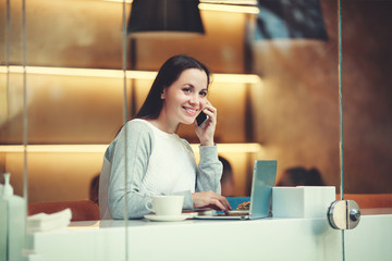 Beautiful Caucasian woman uses Phone while sitting with portable net-book in modern cafe bar, young charming female freelancer thinking about new ideas during work on laptop computer