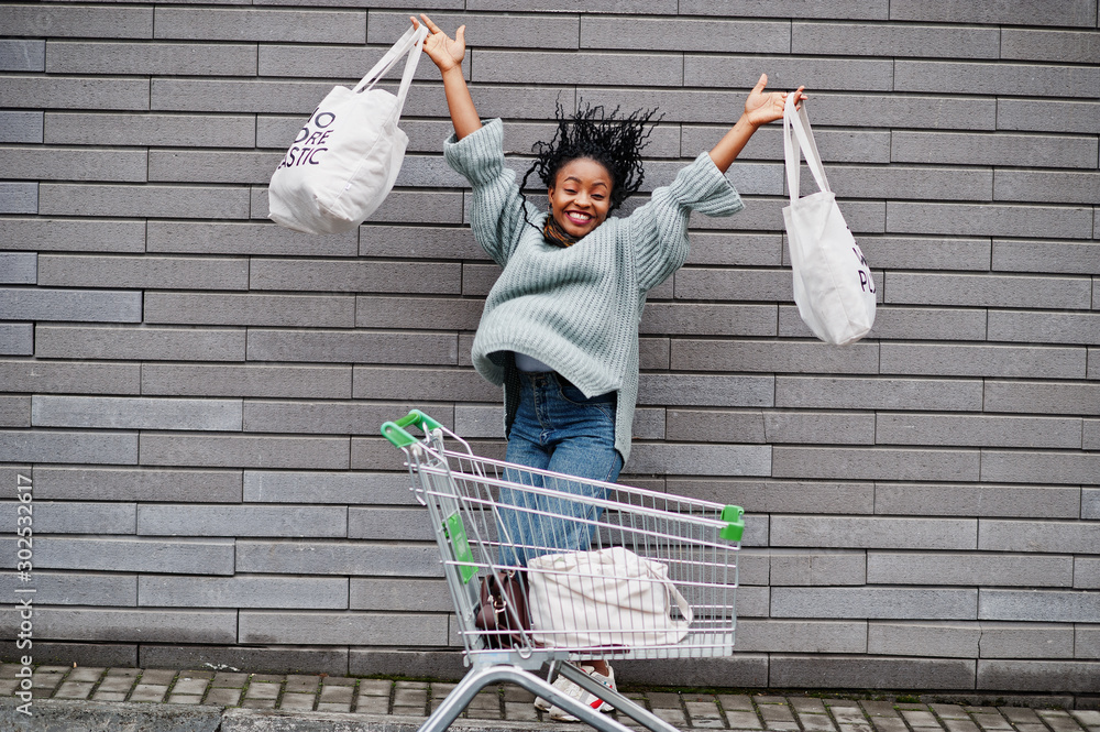 Wall mural No more plastic. African woman with shopping cart trolley and eco bags jump outdoor market.