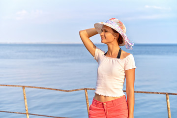 Sideways shot of young woman in summer clothing enjoys picturesque view and oceanscpae in resort town, walks on beach alone, has pleasant warm smile, looks at place of your advertisement, blank form