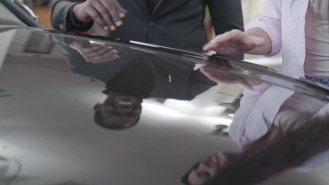 View of new black car hood with reflection of two people. Young Caucasian woman caressing automobile, smiling African American man taking her hand. Multiracial couple admiring new car in showroom.