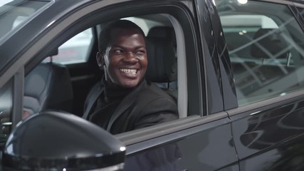 Portrait of happy African American man sitting in vehicle salon and taking keys from Caucasian car dealer. Successful businessman purchasing new automobile in showroom. Car dealership, car business.