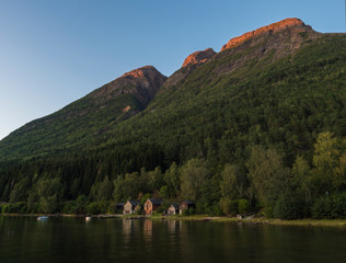View of traditional wooden cottage along Hardanger Fjord in Kinsarvik in Norway with forest rocky hill, fishing boats and reflection in bay. Norwegian landscape, golden hour light and red enlighten