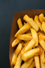 Close up of the golden coloured French fries in a paper box on a dark background.