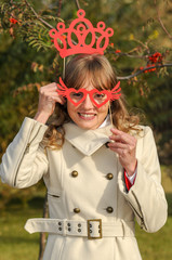 Young beautiful girl with a charming smile and brown hair holds a mask in his hands with glasses and a crown.