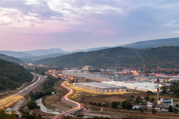 View from above of a large, modern, lighten factory, old, ruined factory, curvy road with bridge and car trails