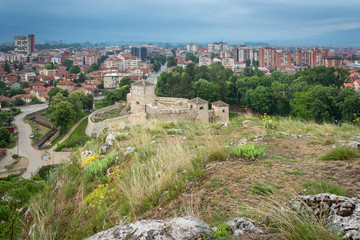 Obraz na płótnie Canvas Low angle shot with foreground rocks and vivid yellow flowers in a foreground and ancient fortress and Pirot cityscape at the distance during blue hour