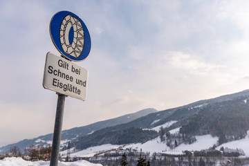 Chain traffic sign: Bei Schnee und Eisglätte. Blue warning information:  Snow chain obligation. Winter time, snow covered road, trees. Styria, Austria