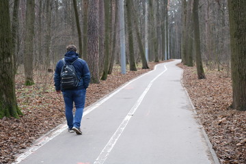 A man in blue clothes with a black backpack walks down a winding asphalt road in an autumn Park on a cloudy day. 