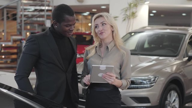 Adult Caucasian woman with blond hair holding tablet and explaining specifications to African American man. Female car dealer standing with customer next to automobile. Car dealership, car business.