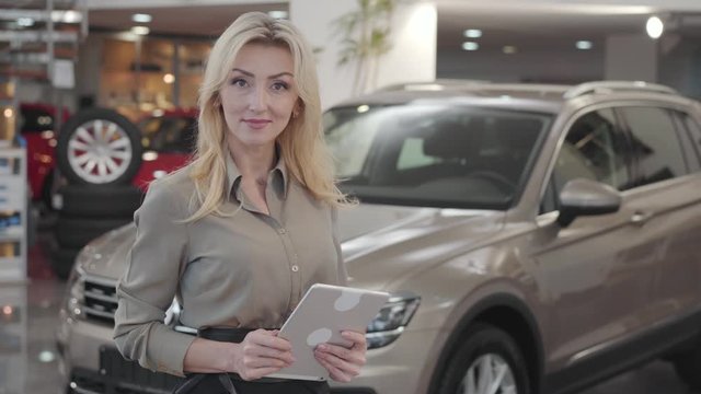 Confident young Caucasian woman using tablet, looking at camera and smiling. Successful blond car dealer posing at the background of new automobiles. Car dealership, car business.