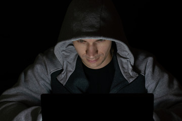 A man with a hood on head stares intently at a computer screen, sitting in the dark