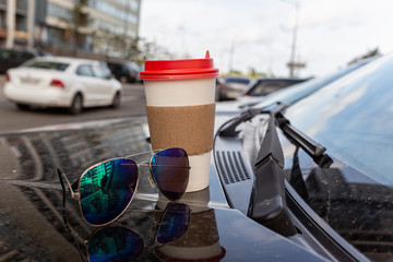 White paper Cup of coffee with the red cover on the hood of the car. Paper Cup with hot tea on the hood of the SUV, closeup on the background of the windshield