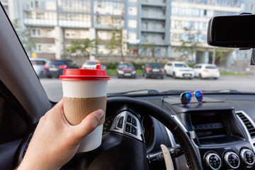 Closeup of a young man car driver drinking coffee, hand holding a paper white coffee Cup with a red lid in the background steering car dashboard blurred Parking background.