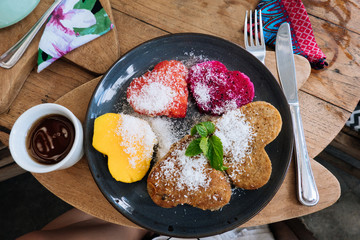 Heart shaped pancakes and tropical fruits on a black plate with mint and honey on wooden table. Valentine's Day. Healthy food concept. Top view.