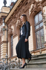 A woman walking down stairs, near old building, in elegant outfit, looking at side, in black leather jacket and high heels. Vertical view.