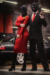 a classic in black costume with a sleeveless jacket and a maid of honor in a mask with weapons standing in a parking lot machine - obrazy, fototapety, plakaty