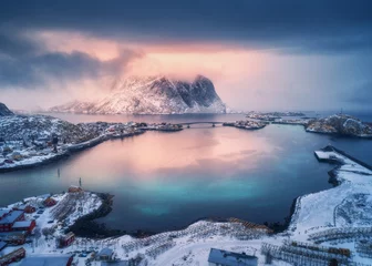  Luchtfoto van besneeuwde berg, dorp aan de zeekust, oranje lucht bij zonsondergang in de winter. Bovenaanzicht van Reine, Lofoten eilanden, Noorwegen. Humeurig landschap met hoge rotsen, huizen, rorbu, weerspiegeling in water © den-belitsky