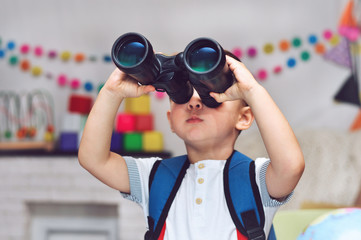 A little boy with a backpack on his back holds binoculars and looks through it.