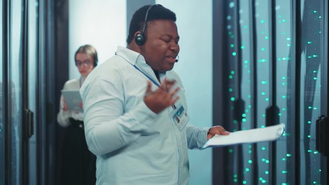Frustrated angry black IT support technician arguing talking on headphones getting bad of difficult working tasks in server rack room. Data center. Stress work.