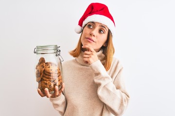 Beautiful redhead woman wearing christmas hat holding cookies jar over isolated background serious face thinking about question, very confused idea