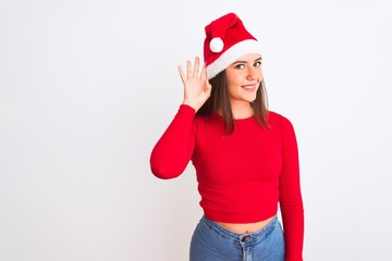 Young beautiful girl wearing Christmas Santa hat standing over isolated white background smiling with hand over ear listening an hearing to rumor or gossip. Deafness concept.