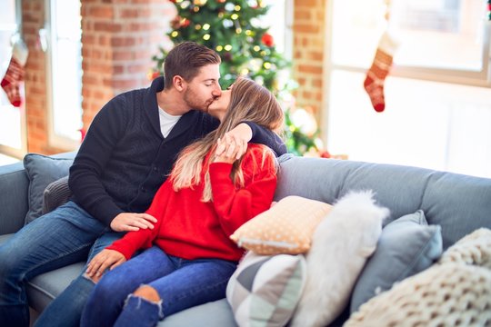 Young beautiful couple smiling happy and confident. Sitting on the sofa hugging and kissing around christmas tree at home