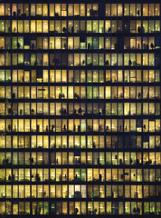 Office building with glass walls and windows illuminated at night. People sitting at the desk, clothes hangers and other objects in silhouette.
