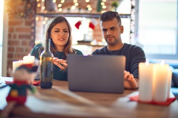 Young beautiful couple sitting using laptop around christmas decoration at home disgusted expression, displeased and fearful doing disgust face because aversion reaction. With hands raised